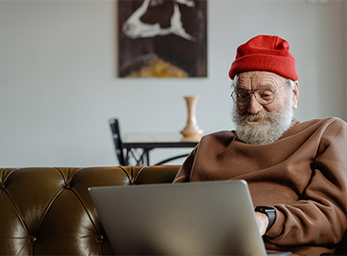 Harmaapartainen mies punainen pipo päässä istuu kannettavan tietokoneen kanssa sohvalla hoitamassa asioita