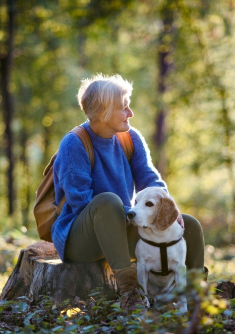 Iäkkäämpi nainen istuu metsässä kannolla koiransa kanssa.